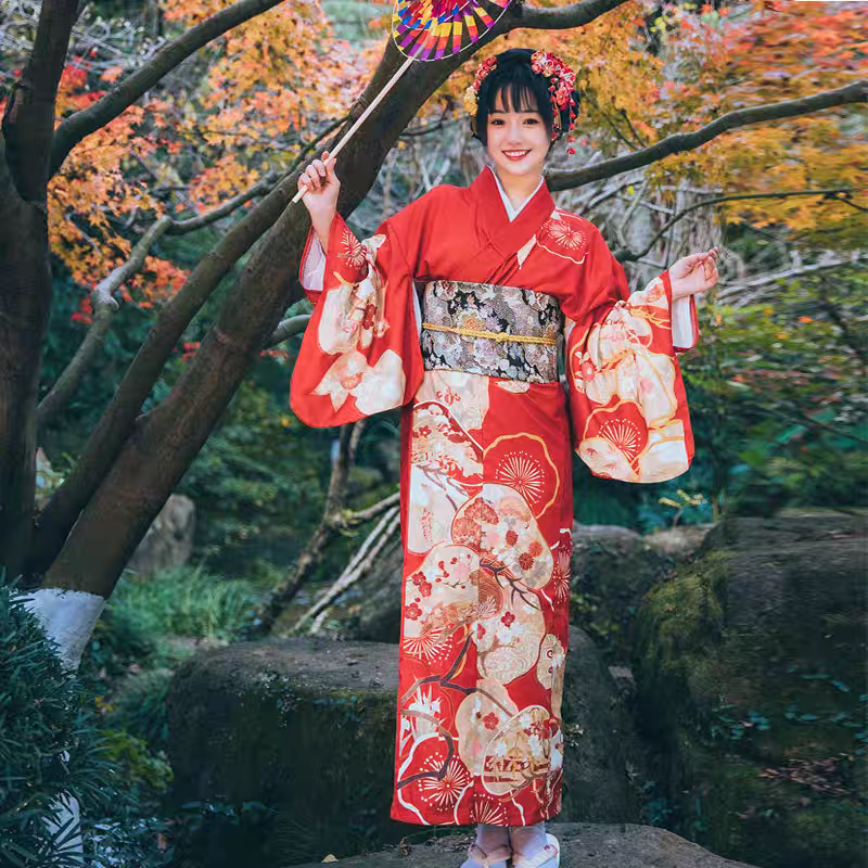 Woman in a traditional red kimono with floral patterns standing in a natural setting with trees and rocks.