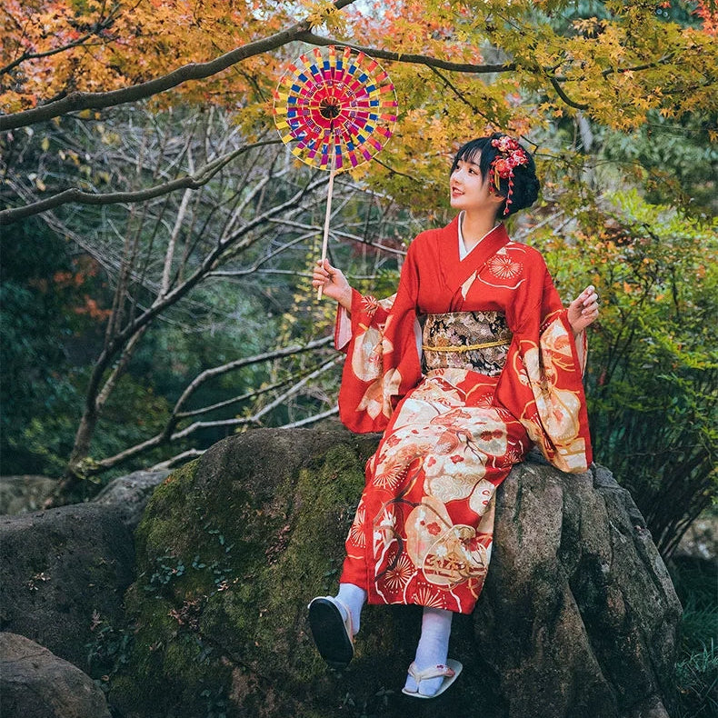 Woman in a red kimono holding a colorful fan in an autumn forest.